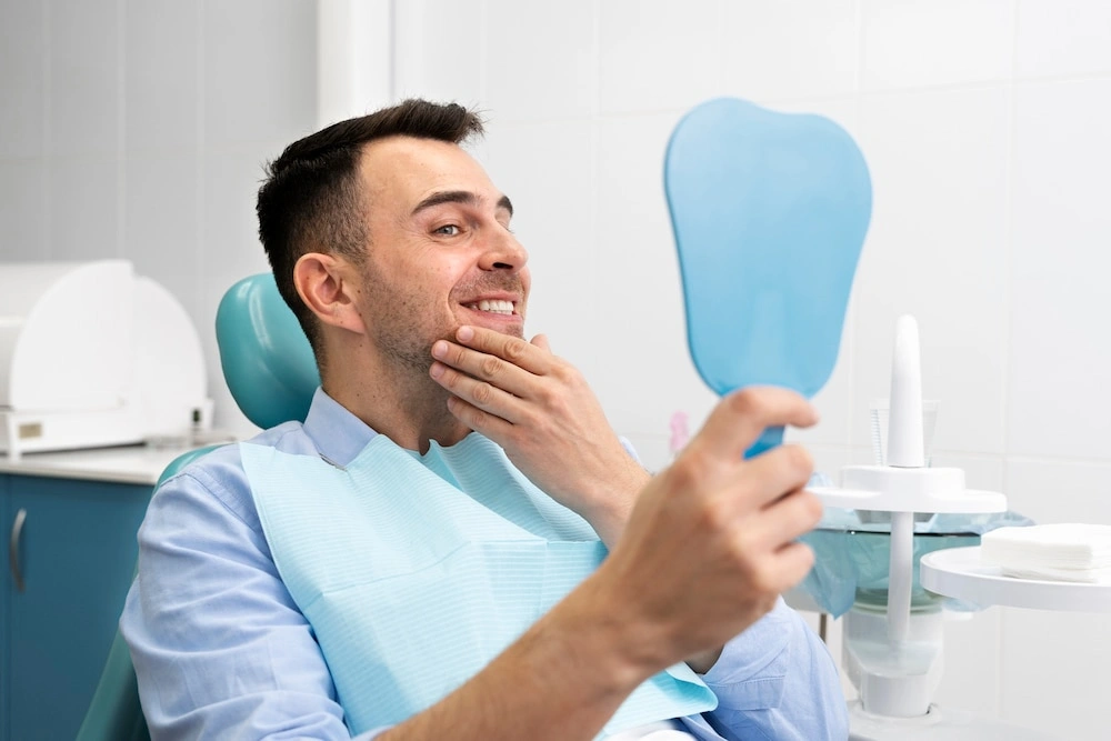 A happy male patient looking in a hand mirror and smiling after receiving dental implants in a clinic.
